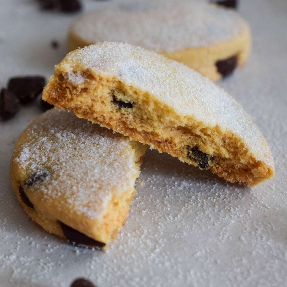 A dark chocolate chip shortbread cookie split in two  with one half sitting on top of the other with a dusting of sugar on them,  on a white background.