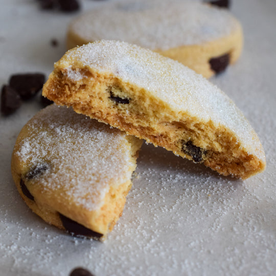 A gluten free dark chocolate chip cookie split in two with one half sitting on top of the other with a dusting of sugar on them, on a white background.