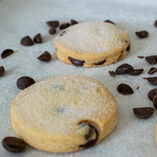 Two dark chocolate shortbread cookies with a dusting of sugar on them. With dark chocolate chips scattered around them. On a white background.