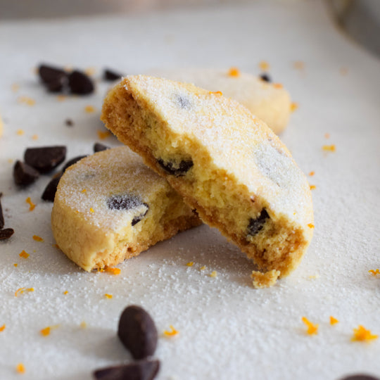 A dark chocolate orange shortbread cookie broken in half with one half leaning up against the other, with a few chocolate chips and orange zest around it on a white background.