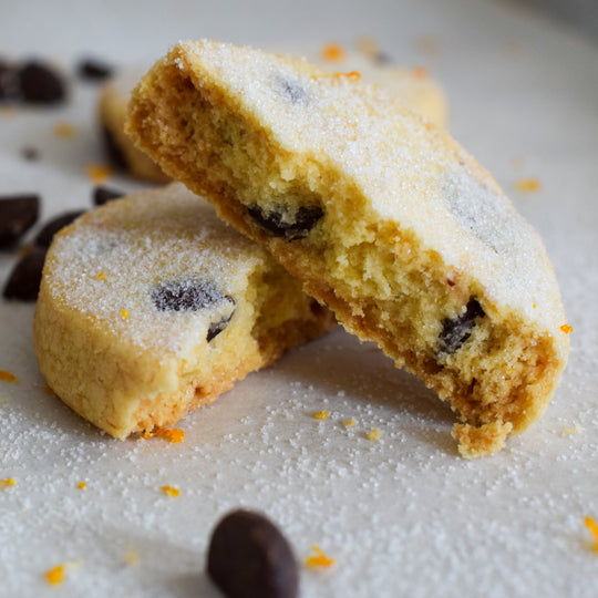 A large dark chocolate orange shortbread cookie broken in half with one half leaning up against the other, with a few chocolate chips and orange zest around it on a white background.