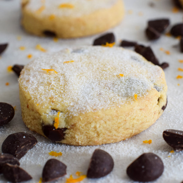 A photo showing dark chocolate orange shortbread cookies, dusted with sugar, on a surface with chocolate pieces and orange slices scattered around.