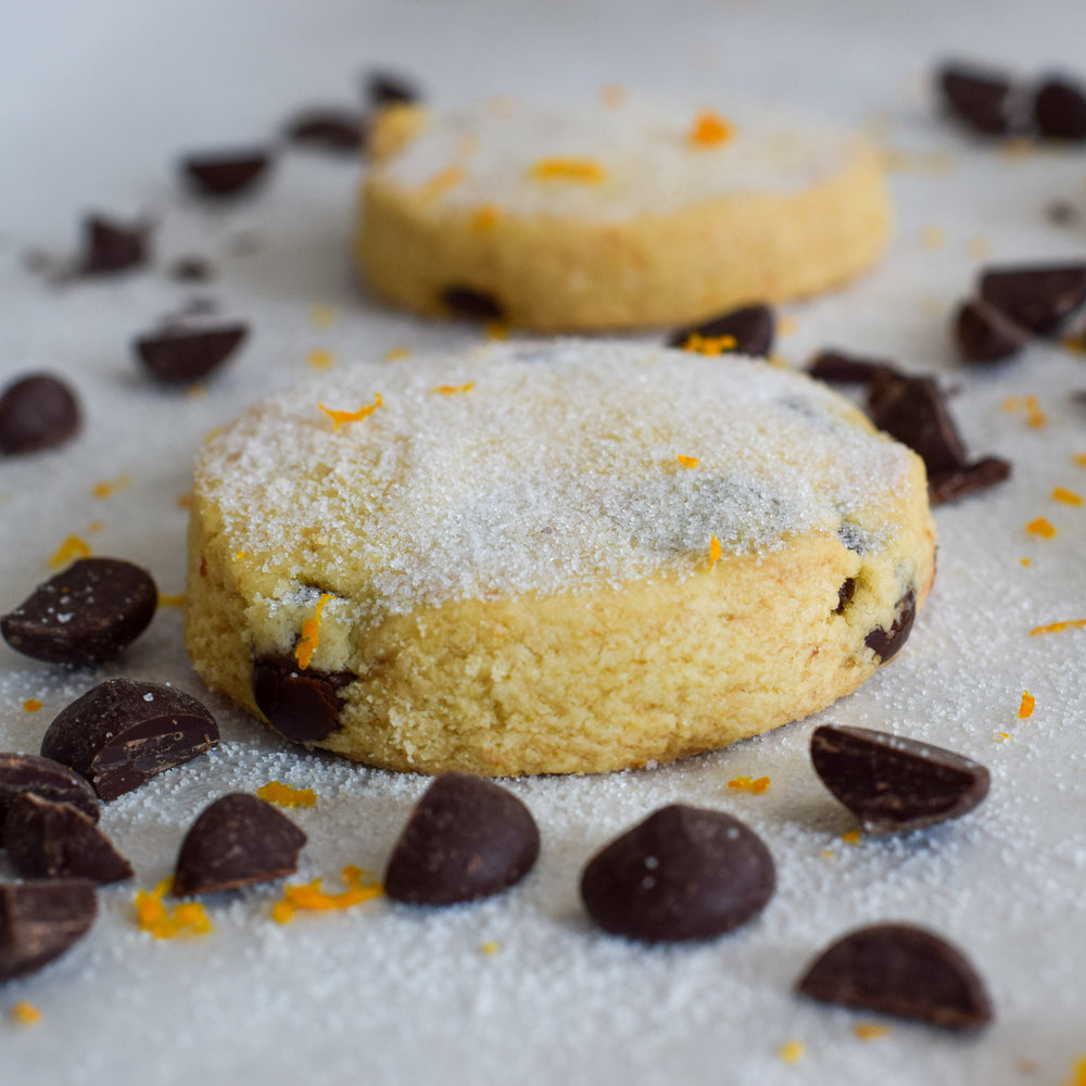 One  large dark chocolate and orange shortbread cookie and one behind it with chocolate chips placed around it with bright orange zest, on a white background.
