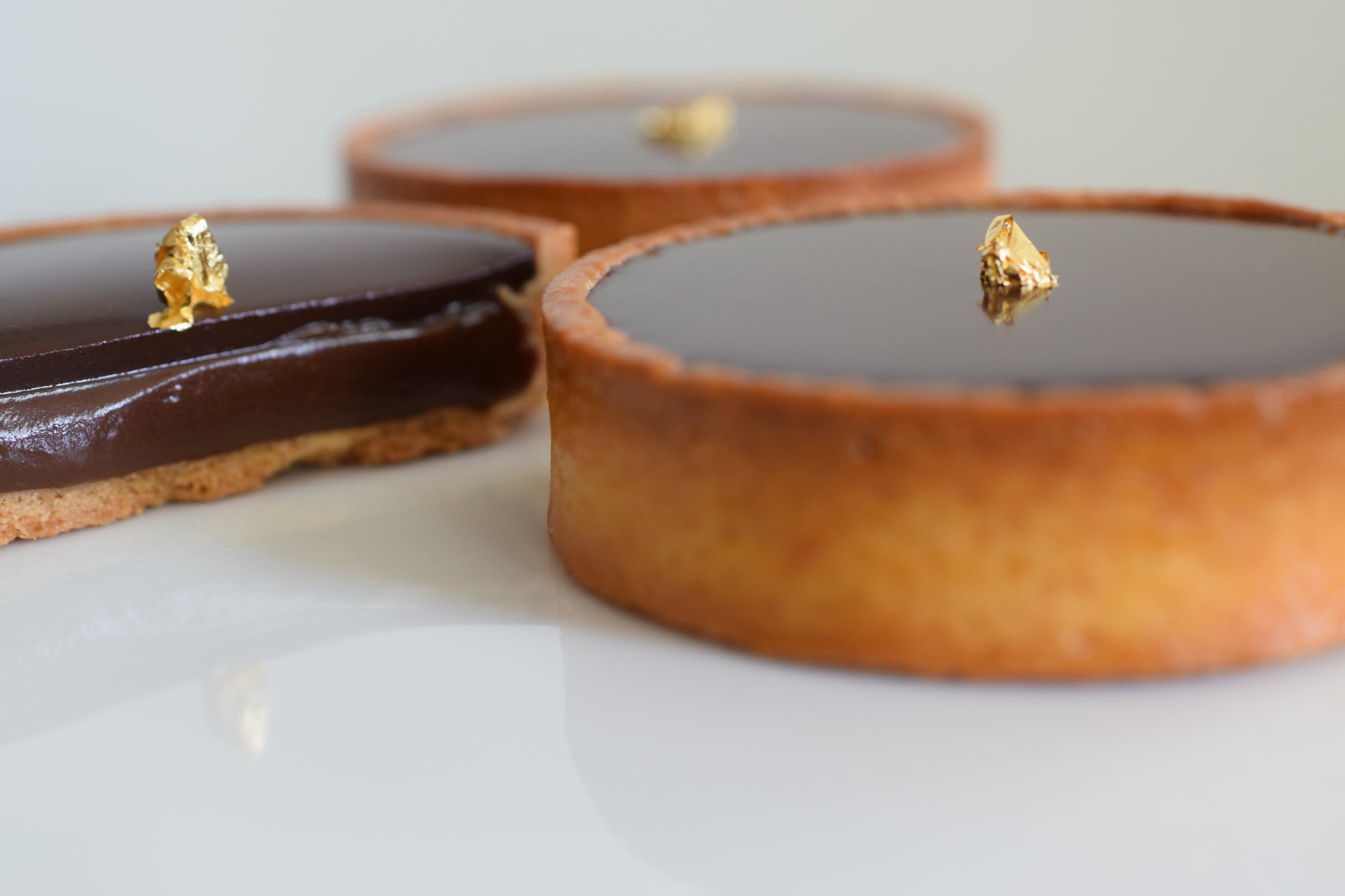 Three chocolate tartlets with glossy dark chocolate ganache and a small gold leaf decoration, arranged on a white surface with a soft-focus background.
