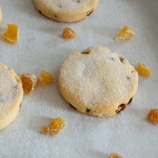 Three gluten free stem ginger shortbread cookie with a dusting of sugar on top of them, with a few pieces of stem ginger around the shortbread cookies. On a white background.