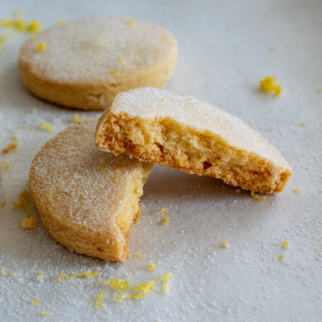 A meyer lemon cookie split in two with one half sitting on top of the other with a dusting of sugar and lemon zest on them, on a white background.