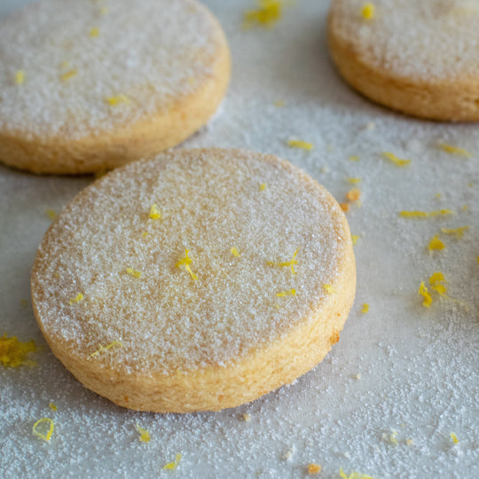 Three gluten free meyer lemon shortbread cookies with a dusting of sugar and lemon zest, on a white background.