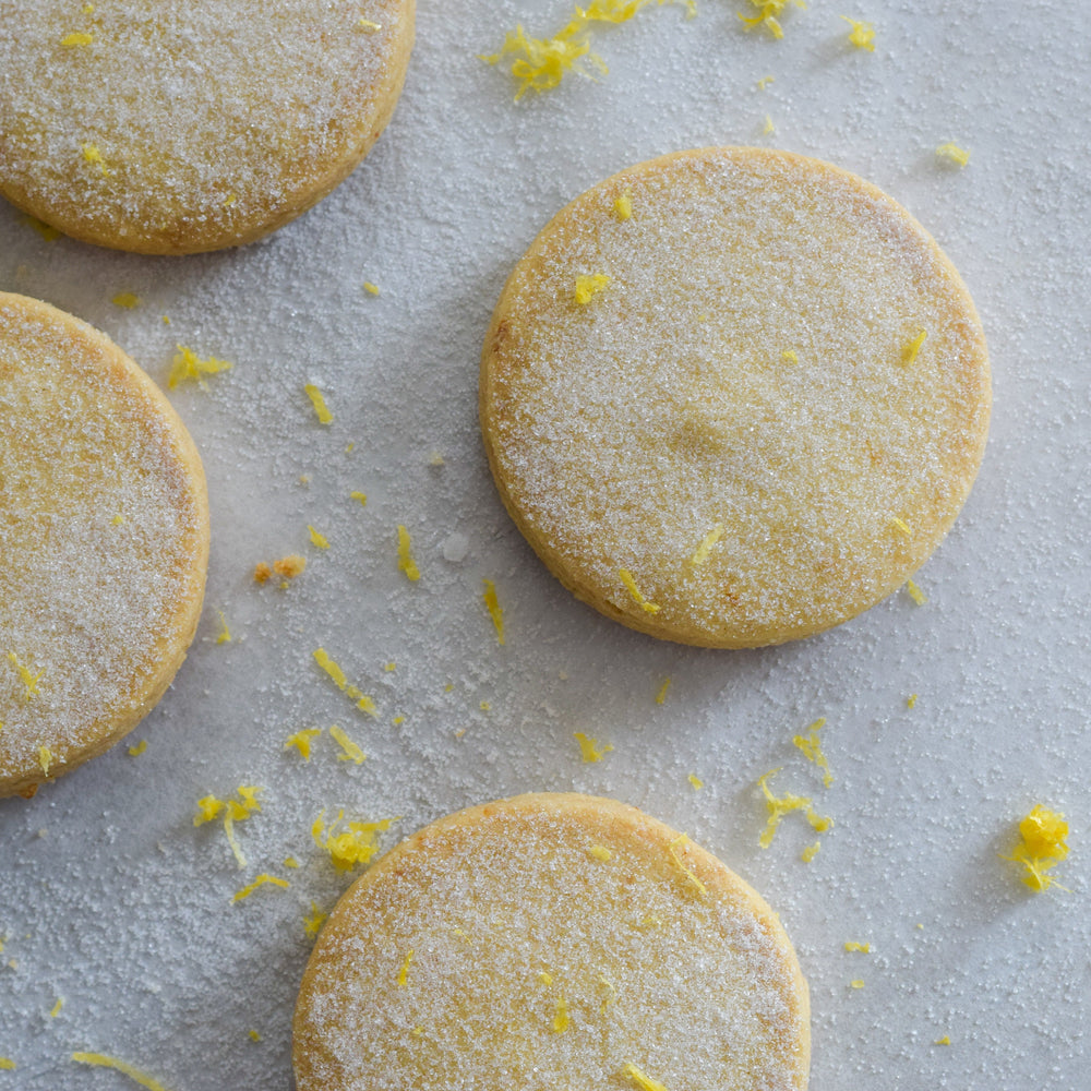 Four meyer lemon shortbread cookies with a dusting of sugar and lemon zest, on a white background.