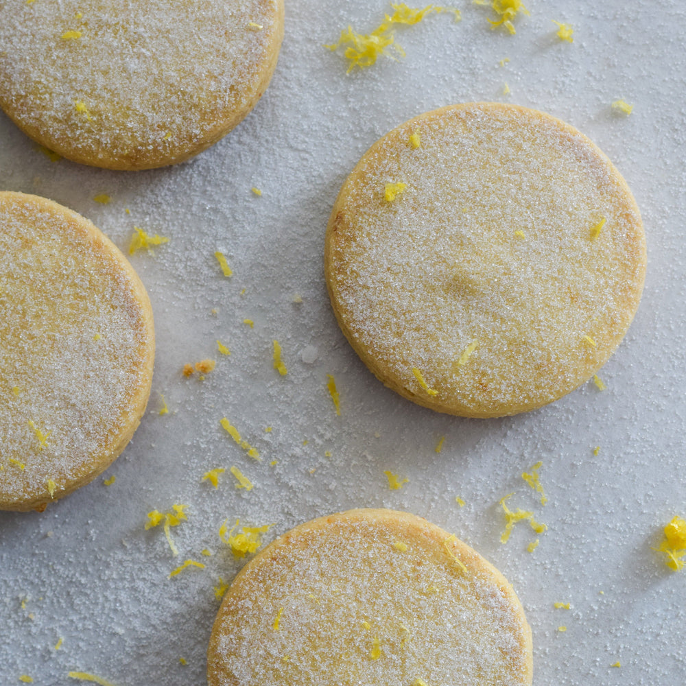 Four gluten free meyer lemon shortbread cookies with a dusting of sugar and lemon zest, on a white background.