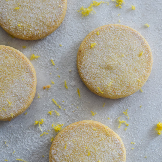 Four gluten free meyer lemon shortbread cookies with a dusting of sugar and lemon zest, on a white background.