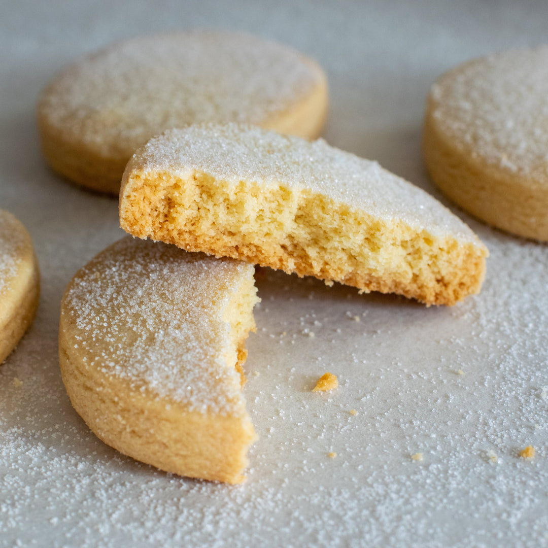 A shortbread cookie broken in half with one half leaning up against the other with a few crumbs and a dusting of sugar in the foreground. Three more cookies behind it on a white background.