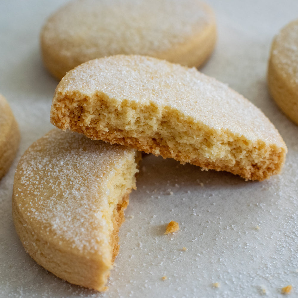 A shortbread cookie broken in half with one half leaning up against the other with a few crumbs and a dusting of sugar in the foreground. Three more cookies behind it on a white background.